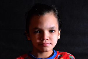 Portrait of a young child with dark hair, wearing a red shirt with colorful patterns, against a dark background.